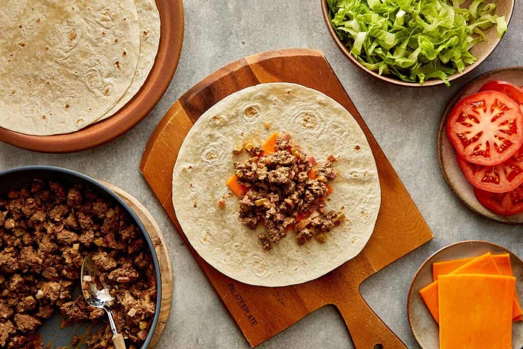 a top down view of a wrap on top of a wooden board sitting on a kitchen bench, the wrap has a slice of cheese and some cooked mince on it - 11