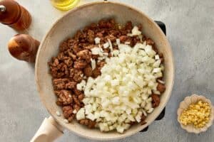 Browned ground beef with diced onion in a pan, with minced garlic on the side ready to be added