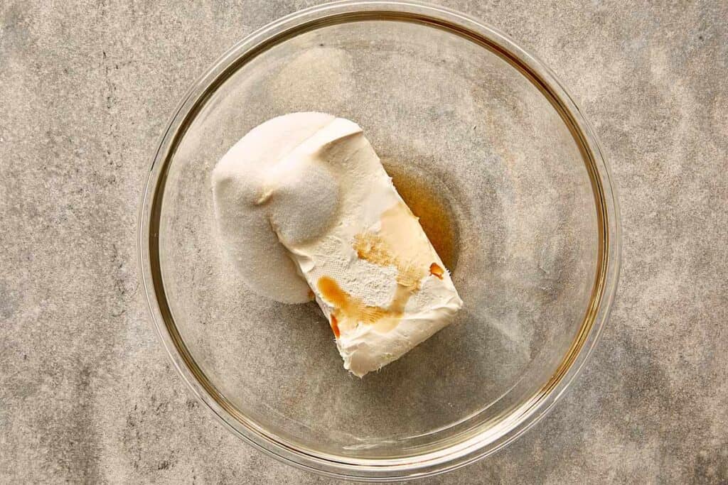 A top down view of a glass bowl with softened cream cheese and sugar in the bottom and what looks like vanilla essence on top pooling in the bottom as well