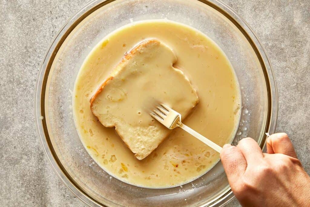 a slice of bread being soaked in a mix of egg and milk in a glass bowl on top of a kitchen bench