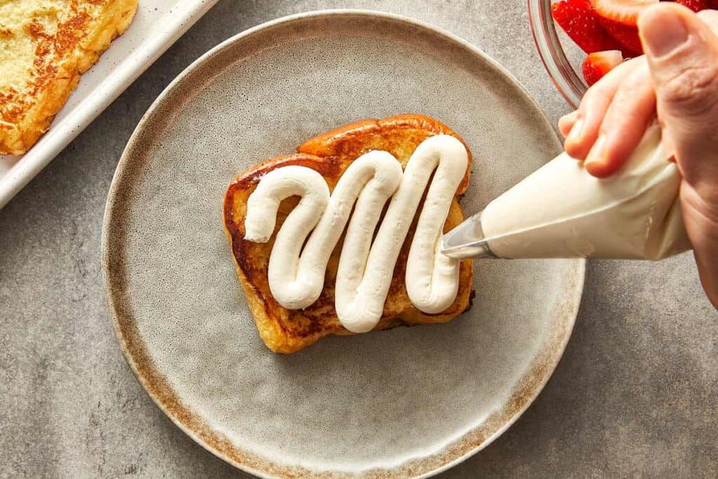 a top down shot of a brioche toast sitting on a hand made plate being covered with a piping bag frosting in a zig zag on top.