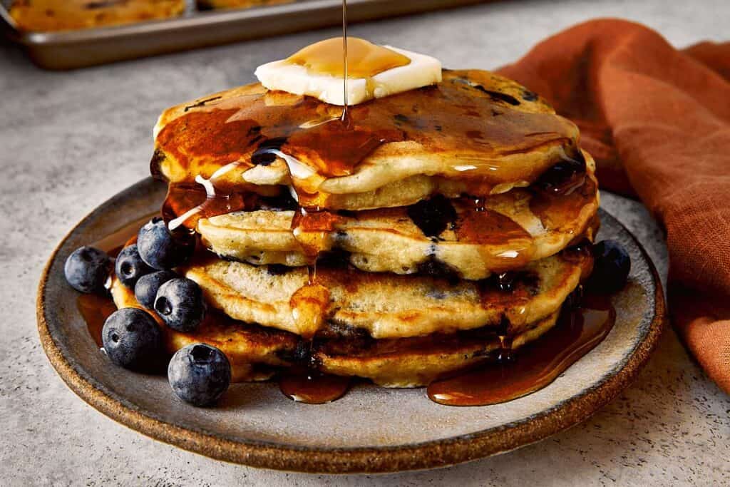 Fluffy blueberry chocolate chip pancakes stacked on a plate, topped with butter and warm maple syrup being poured over, with fresh blueberries on the side.