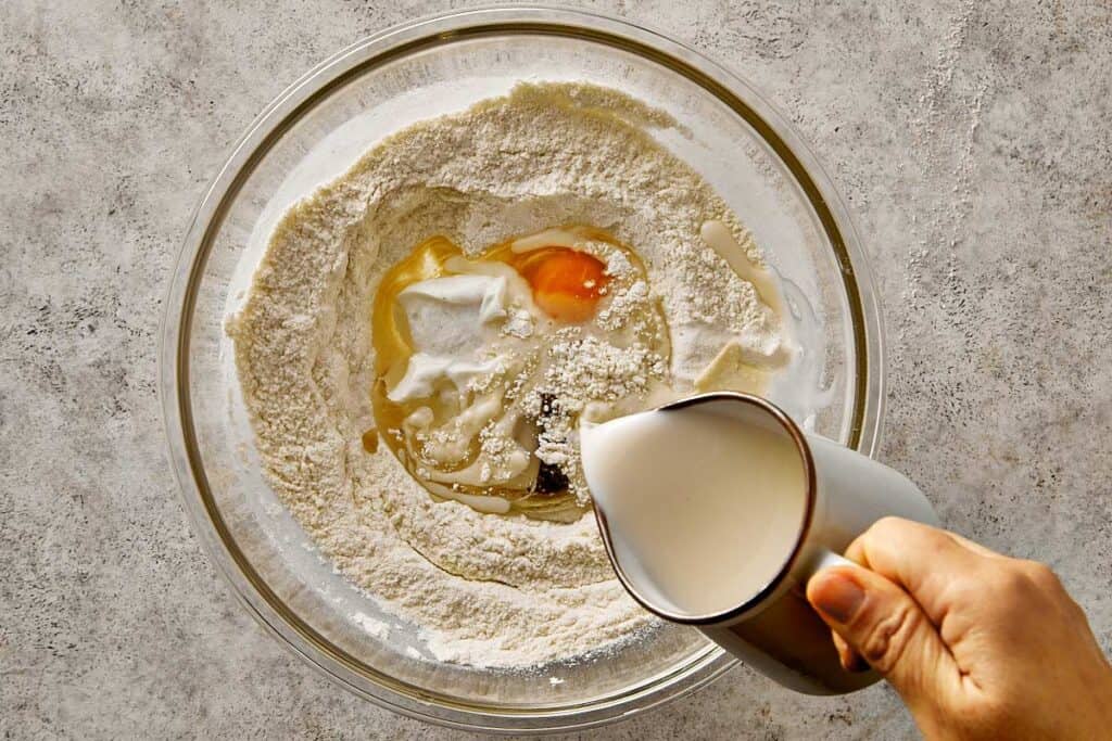 Adding wet ingredients to the dry mixture for blueberry chocolate chip pancakes, including egg, Greek yogurt, vanilla, coconut oil, and almond milk in a mixing bowl.