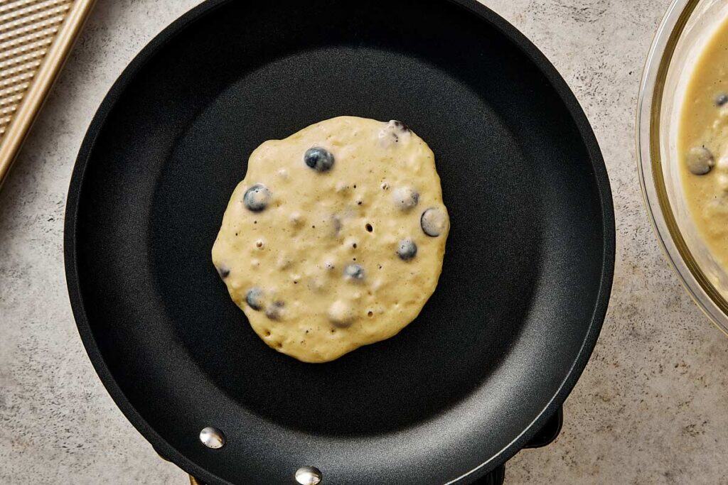 Blueberry chocolate chip pancake batter cooking on a nonstick skillet, showing bubbles forming on the surface before flipping.