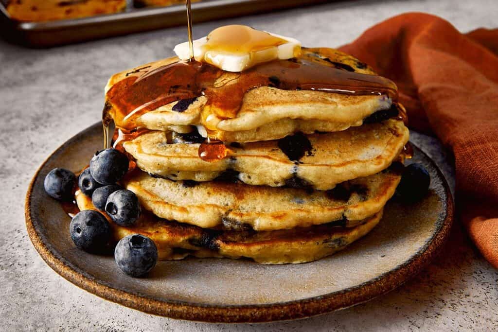 Stack of fluffy blueberry chocolate chip pancakes topped with butter and maple syrup, served on a plate with fresh blueberries on the side.