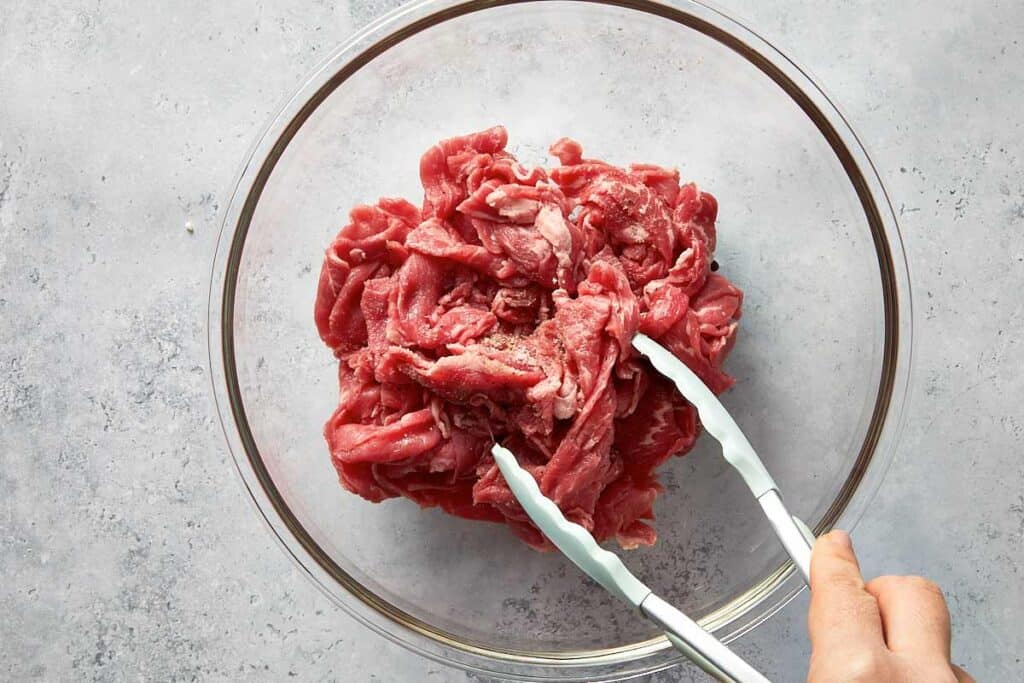 Thinly sliced raw steak seasoned with salt and pepper in a glass bowl, being tossed with tongs before cooking.