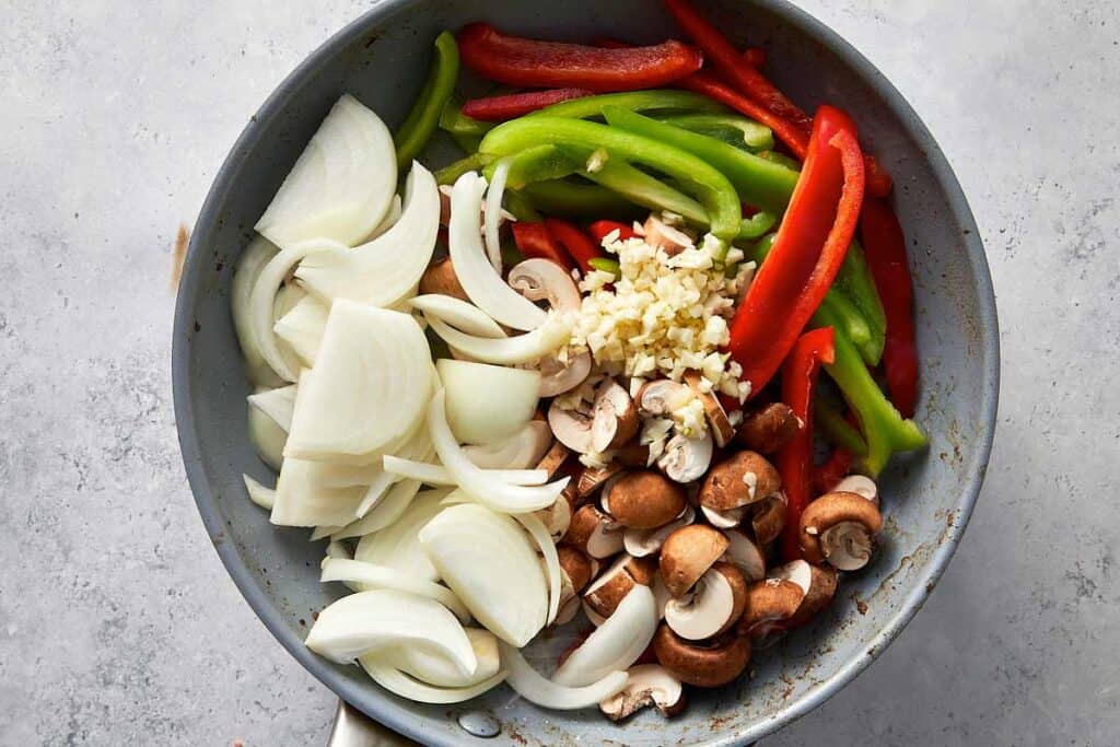 Sliced onions, red and green bell peppers, mushrooms, and minced garlic added to the same skillet to sauté until tender and aromatic.