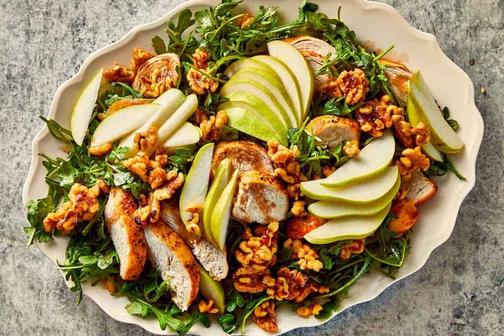 Arugula salad with grilled chicken, sliced pears, and caramelized walnuts arranged on a serving platter.