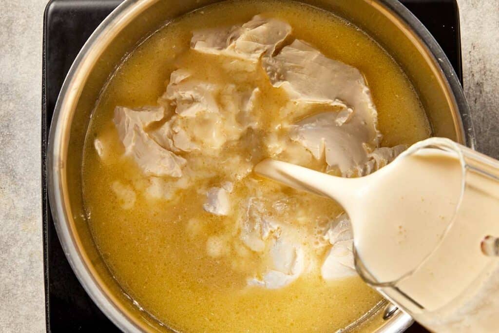 Light cream being poured into a pan with cream cheese and chicken stock to create a smooth base for creamy corn