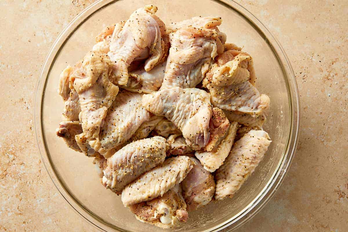 Raw chicken wings tossed with lemon pepper seasoning and baking powder in a bowl, evenly coated before cooking