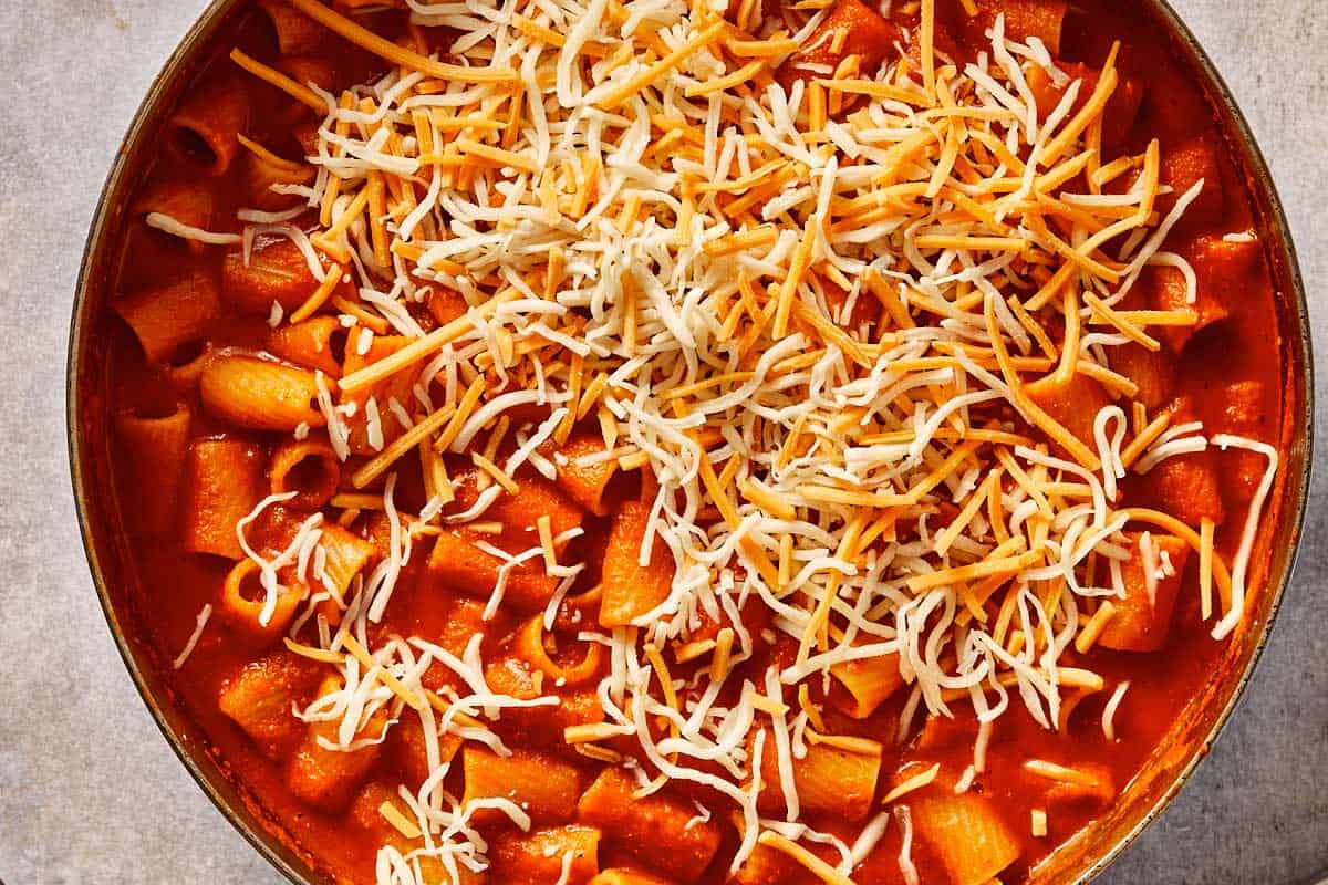 Cooked rigatoni in tomato soup topped with shredded cheddar and mozzarella cheese, ready to be baked until melted and golden.