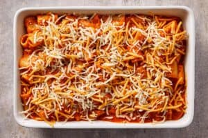 Tomato pasta mixture transferred to a baking dish and topped with shredded cheddar and mozzarella cheese before baking.