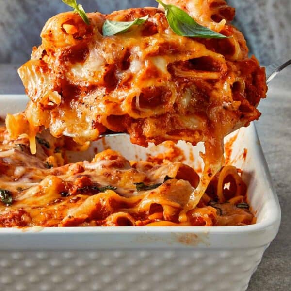 Close-up of tomato soup mac and cheese being served, with melted cheese stretching over rigatoni pasta in a rich tomato sauce, topped with fresh basil.