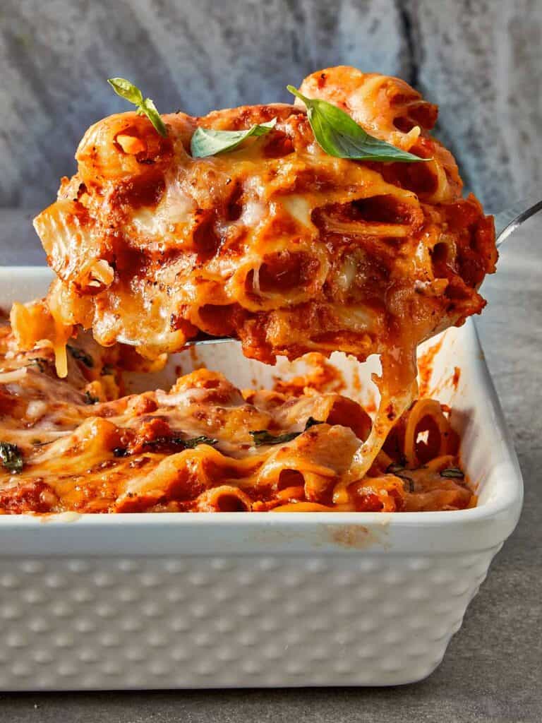 Close-up of tomato soup mac and cheese being served, with melted cheese stretching over rigatoni pasta in a rich tomato sauce, topped with fresh basil.