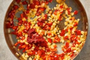 Chopped onion and red bell pepper sautéing in a pan with tomato paste, cooking until soft and fragrant.