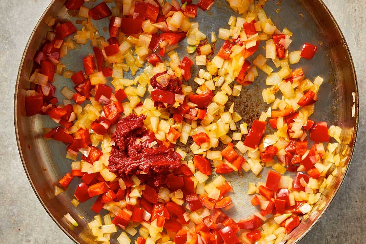 Chopped onion and red bell pepper sautéing in a pan with tomato paste, cooking until soft and fragrant.