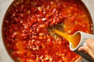 Vegetable broth being poured into sautéed onion, red bell pepper, and tomato paste, creating the base for the tomato soup.