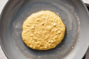 Cottage cheese pancake cooking in a pan, starting to set on the surface with small bubbles forming.