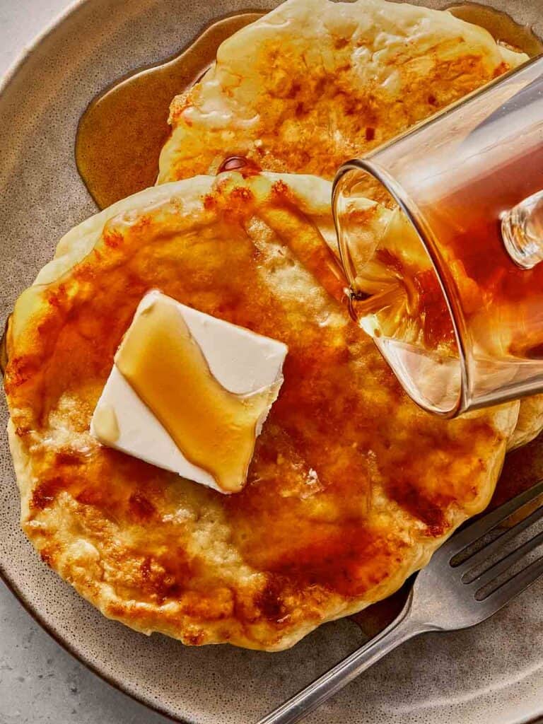Cottage cheese pancakes on a plate with butter melting on top, drizzled with maple syrup as it’s being poured, showing a soft texture and golden surface.