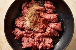Raw ground beef in a skillet seasoned with spices including paprika, garlic powder, and onion powder.