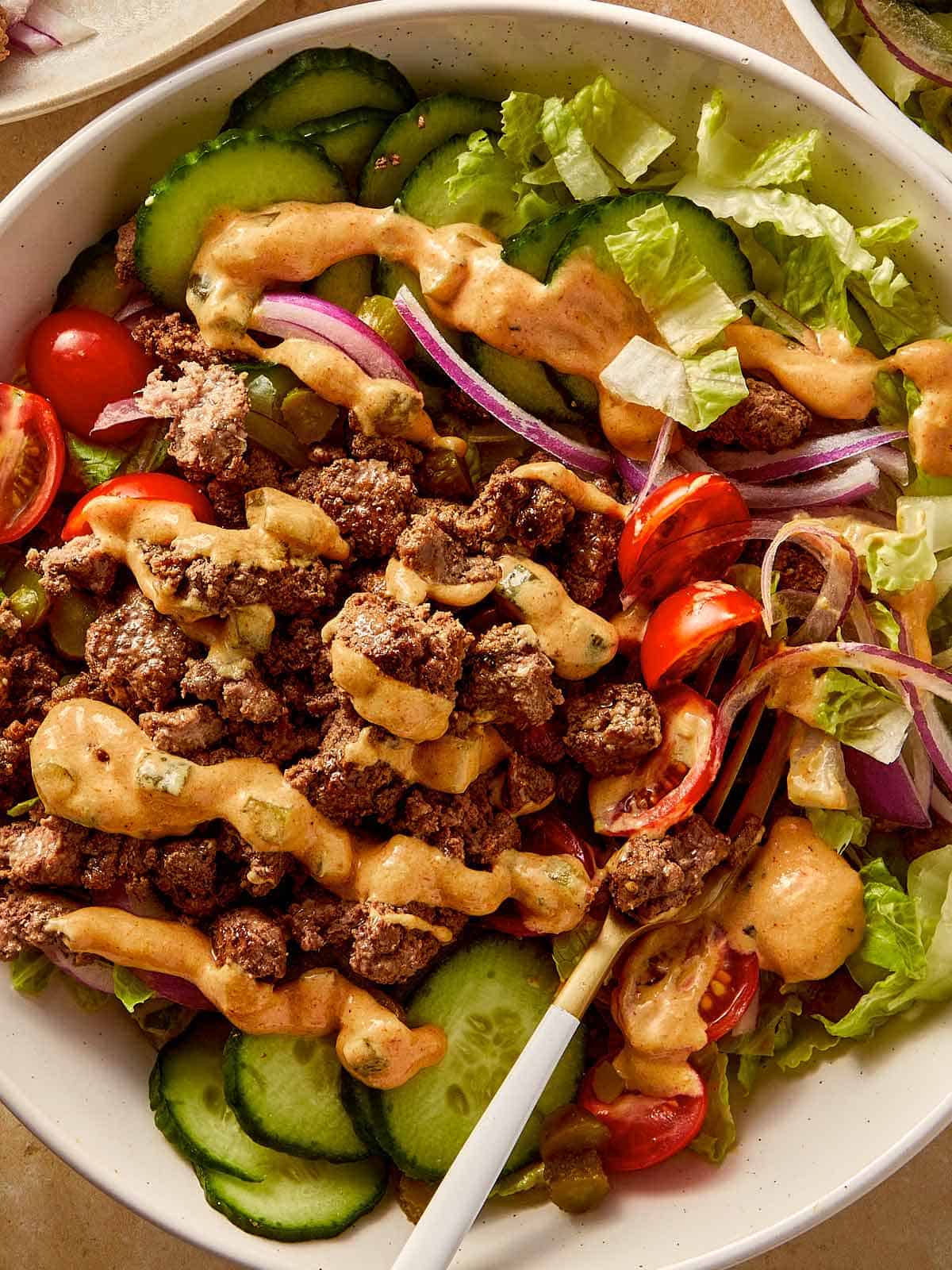Burger bowl with ground beef, cucumber, cherry tomatoes, red onion, lettuce, and creamy burger sauce drizzled on top.