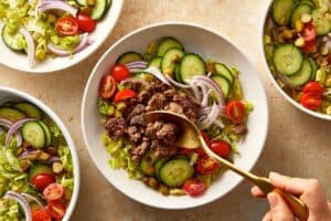Cooked ground beef being added on top of a vegetable-filled burger bowl.