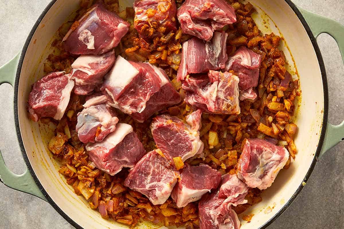 Raw lamb pieces added on top of spiced onions in a pan.