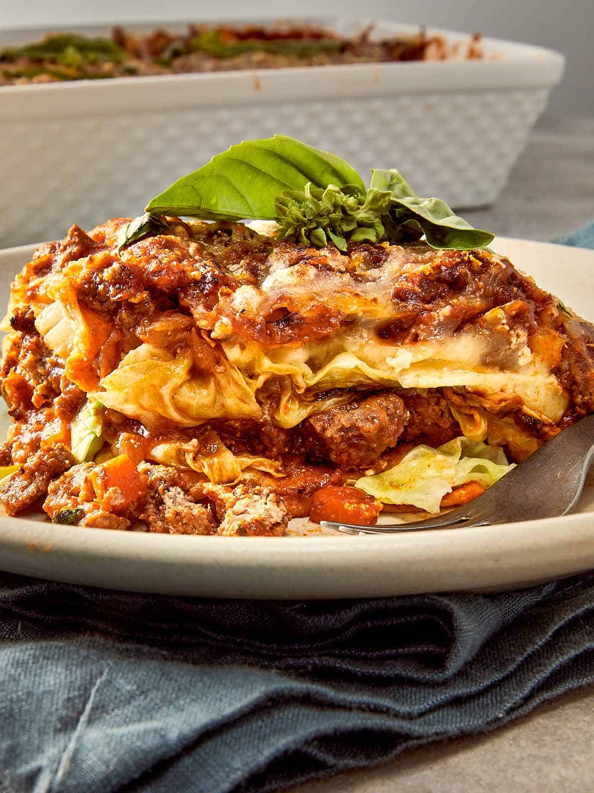 Slice of cabbage ricotta lasagna layered with beef meat sauce, melted mozzarella, ricotta filling, and tender cabbage leaves on a white plate with fresh basil on top.