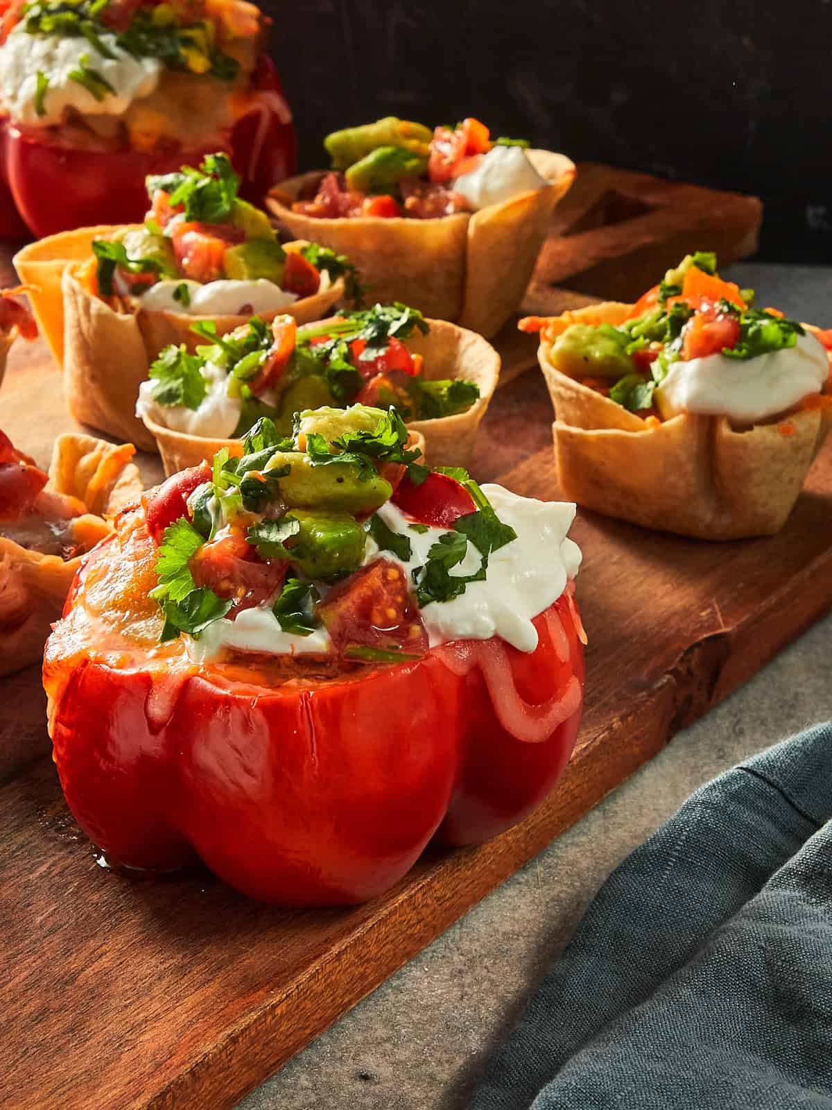 A close-up shot of multiple baked appetizer cups on a wooden serving board, featuring a large, stuffed red capsicum cup in the foreground and several mini tortilla taco cups in the background. All are filled with seasoned chicken, melted mozzarella cheese, and topped generously with sour cream and fresh avocado pico de gallo with coriander.