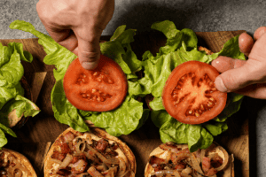A top-down view of hands placing thick tomato slices over fresh green lettuce on the top halves of toasted burger buns.