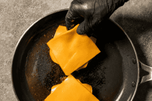 Placing slices of cheddar cheese on top of a juicy homemade beef hamburger patty in a skillet to melt.