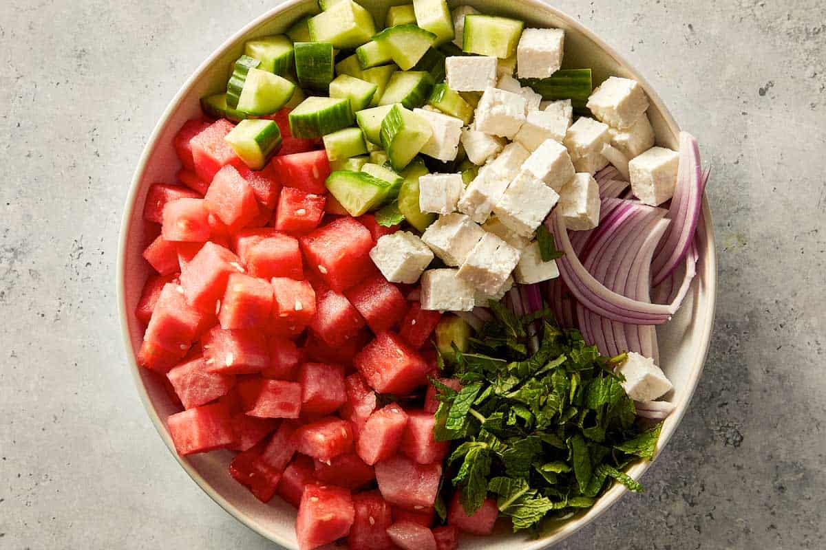 Cubed watermelon, cucumber, Danish feta, sliced red onion, and chopped mint arranged in a large mixing bowl.