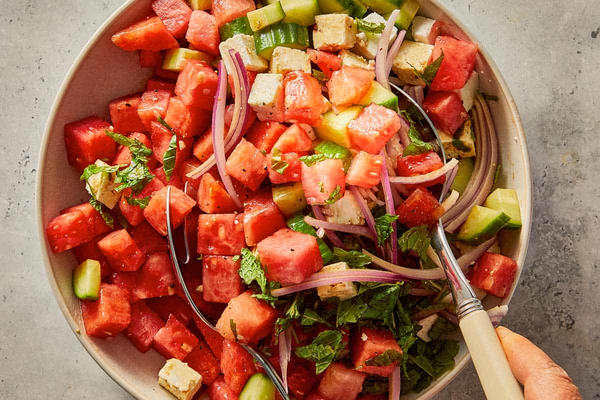 Tossing watermelon feta salad in a large bowl with two serving spoons.