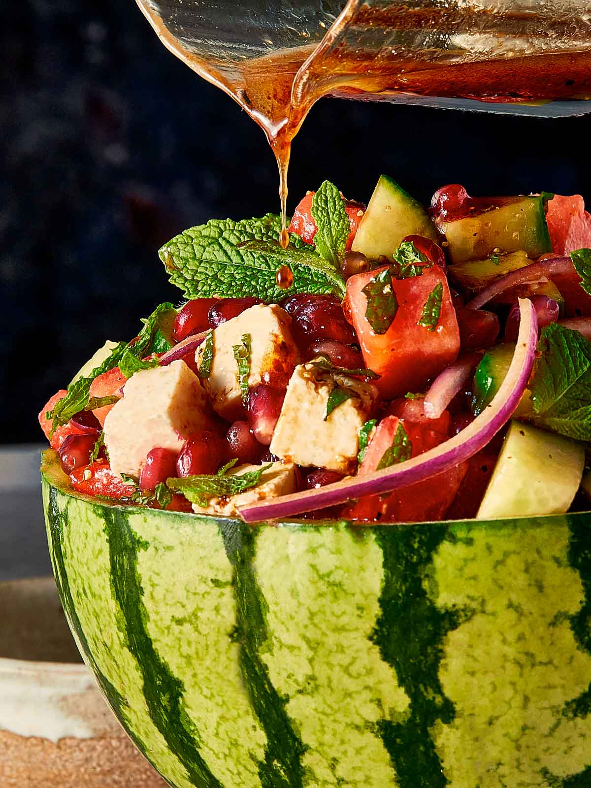 Pouring dark pomegranate molasses dressing over a fresh watermelon and feta salad sitting inside a hollowed out watermelon rind.