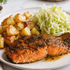 A photo of a sockeye salmon fillet cooked on a white plate, cooked with herbs on top. In the background of the white plate you have cubed potatoes and shredded cabbage salad