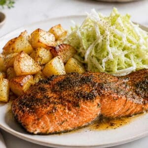 A photo of a sockeye salmon fillet cooked on a white plate, cooked with herbs on top. In the background of the white plate you have cubed potatoes and shredded cabbage salad