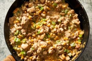 Overhead close-up of a black skillet filled with diced burrito chicken, sweet corn, onions, and green capsicum simmering together in a thick, creamy cheese sauce.