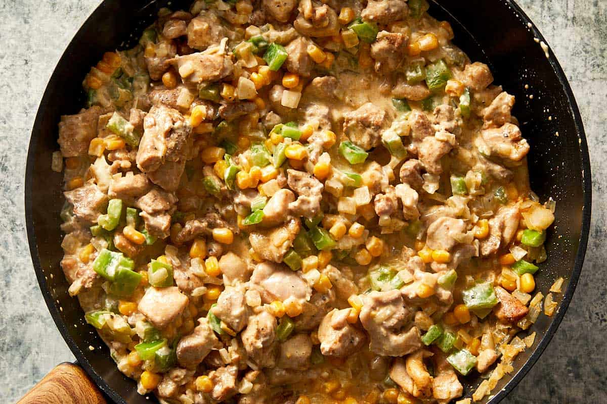 Overhead close-up of a black skillet filled with diced burrito chicken, sweet corn, onions, and green capsicum simmering together in a thick, creamy cheese sauce.