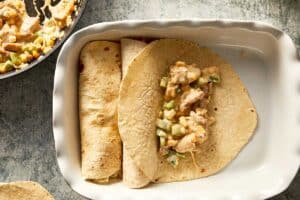 Overhead view of a white baking dish showing the assembly of a chicken burrito, featuring two fully rolled flour tortillas and a third open tortilla topped with a creamy chicken, corn, and green capsicum filling next to a partially visible skillet of the remaining mixture.