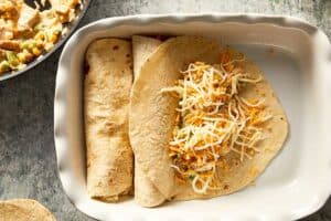 Overhead view of a white baking dish showing two rolled flour tortillas and a third open tortilla topped with a creamy chicken filling and a generous sprinkle of shredded cheese, demonstrating the assembly