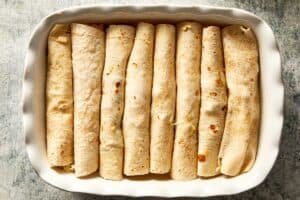 Overhead view of a white baking dish filled with eight tightly rolled flour tortillas arranged closely side-by-side in a single layer.