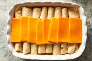 Overhead view of a white baking dish containing eight rolled flour tortillas lined up side-by-side, with overlapping slices of orange cheddar cheese placed across the middle of the rolls.