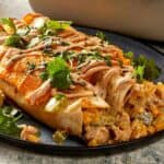 close-up square shot of baked flour tortilla rolls resting on a blue plate. The open ends reveal a creamy chicken, sweet corn, and green capsicum filling, while the tops are covered in melted cheddar cheese, a zigzag drizzle of sauce, and scattered fresh cilantro.