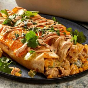 close-up square shot of baked flour tortilla rolls resting on a blue plate. The open ends reveal a creamy chicken, sweet corn, and green capsicum filling, while the tops are covered in melted cheddar cheese, a zigzag drizzle of sauce, and scattered fresh cilantro.