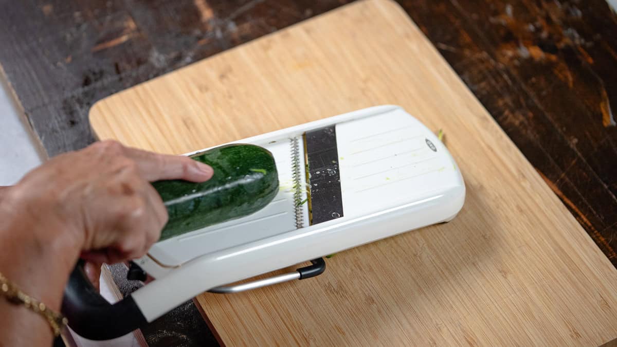 hands using a white mandoline slicer to cut a green zucchini lengthwise on a wooden cutting board.