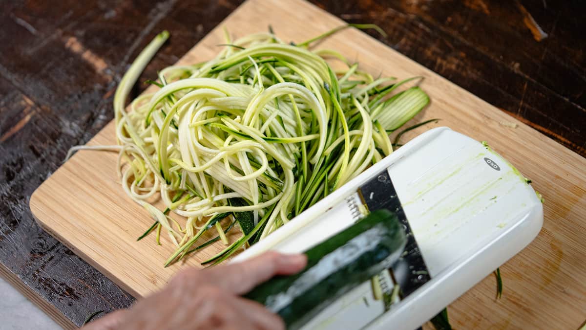 A large pile of thinly sliced, fresh zucchini noodles resting on a wooden cutting board next to a white mandoline slicer.
