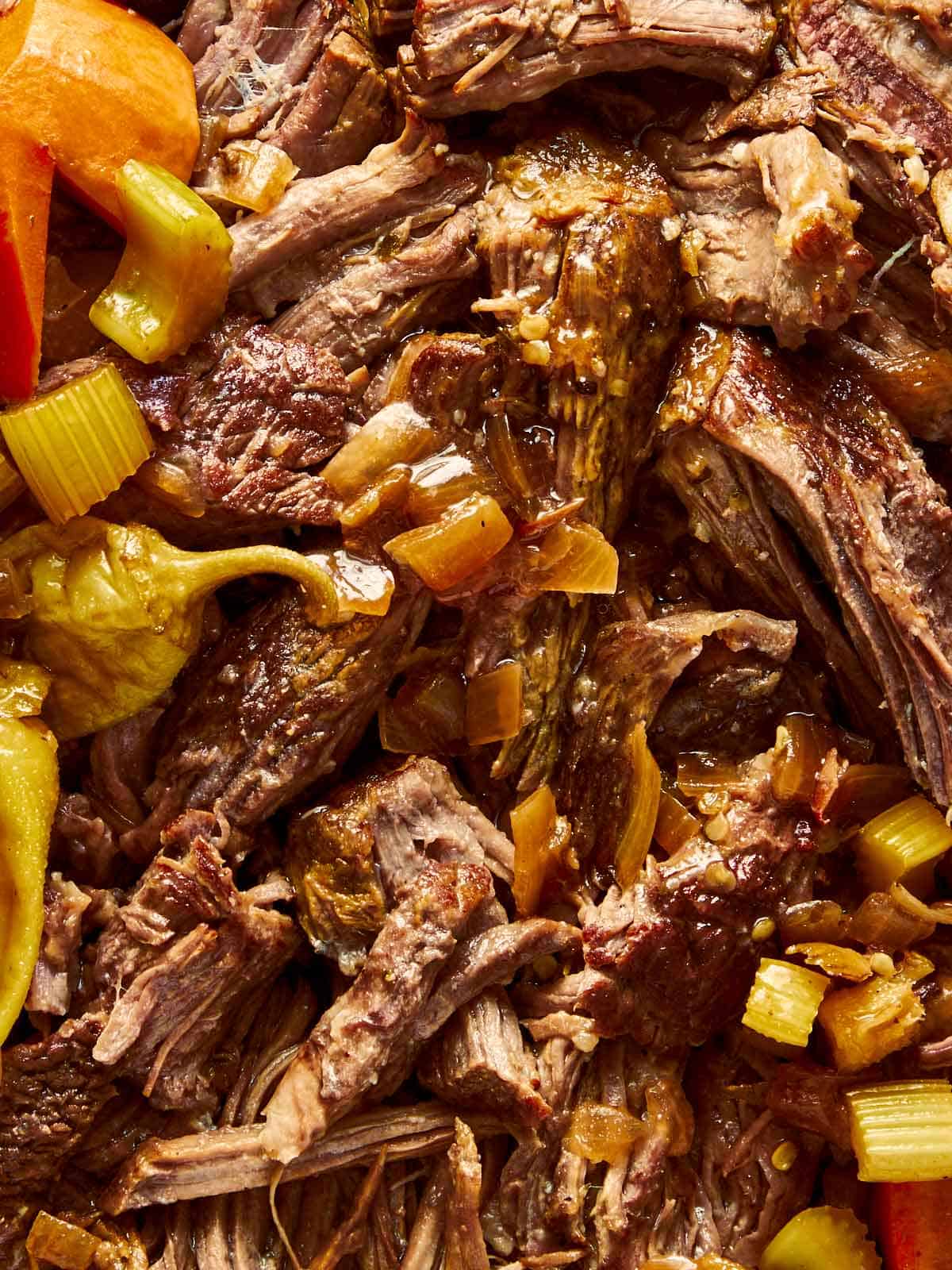 Close-up of shredded Mississippi pot roast in a dark pot with cooked carrots, celery, pepperoncinis, and gravy