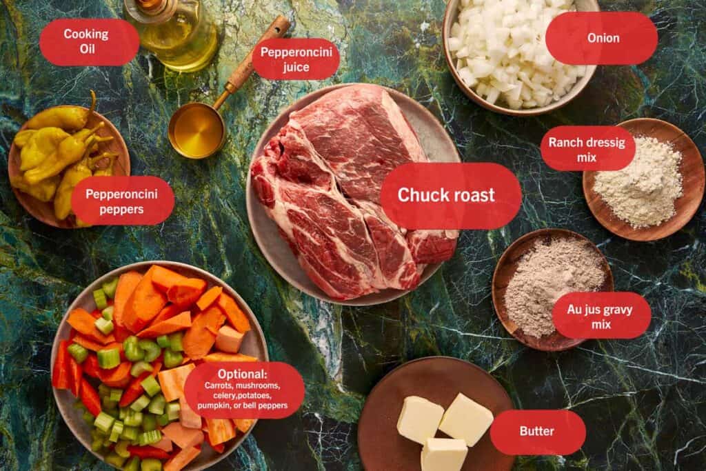 Raw ingredients for Mississippi pot roast laid out on a green marble surface, featuring a large raw chuck roast, diced onions, whole pepperoncinis, chopped carrots and celery, cubed butter, cooking oil, and dry seasoning packets.