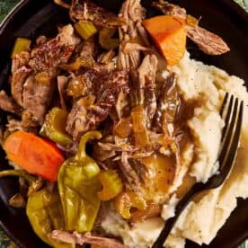 A dark bowl of shredded Mississippi pot roast with gravy, cooked carrots, celery, and whole pepperoncinis, served over creamy mashed potatoes.
