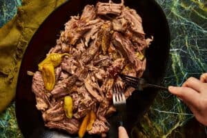 A top-down view of a dark oval platter filled with shredded beef and cooked pepperoncini peppers, as two hands use forks to pull the meat apart on a green marbled surface.